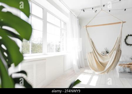 Stilvolles, trendiges Interieur im skandinavischen Stil. Großes Holzfenster sieht weiße Macrame Hängematte vor dem Hintergrund der grünen Monstera, Bett auf der Floo Stockfoto