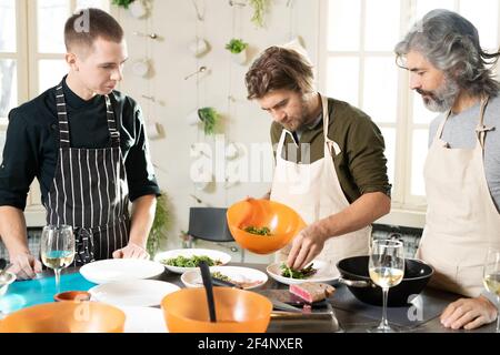 Junger bärtiger Mann in Schürze, der sich über einen großen Küchentisch beugte Und legen frische Ruccola auf Teller unter reifen Lernenden und Kochtrainer Stockfoto