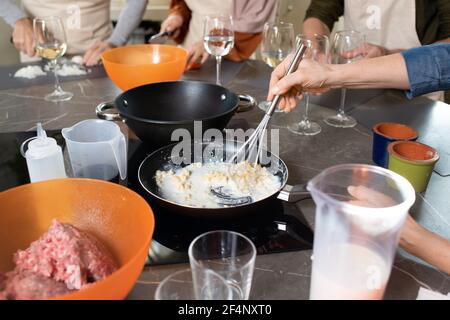 Hände reifer Frau, die Milch und Butter einmischt Bratpfanne gegen andere Auszubildende Kochen neuen Kurs durch große Tisch in der Küche Stockfoto