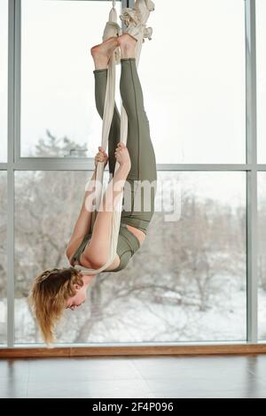 Rückansicht einer aktiven jungen Frau, die Antigravity Yoga macht Im Fitnessstudio, während Sie kopfüber hängen und an Weiß festhalten Hängematte Stockfoto