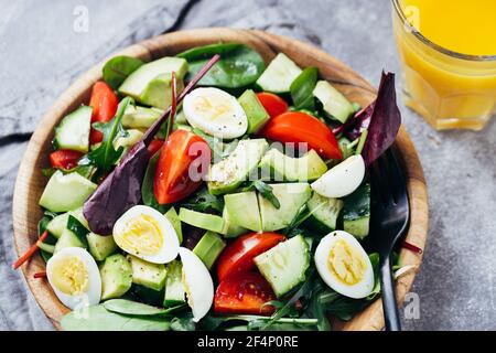 Grüner Salat mit Tomaten, Avocado, Gurken und Eiern auf Holzplatte und Orangensaft auf dem Tisch. Stockfoto