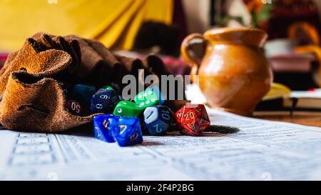 Rollenspiel Würfel in verschiedenen Formen und Farben, die aus einem Lederwürfelbeutel auf einem Charakterblatt auslaufen. Mit im Hintergrund ein Keramik-Trinkglas A Stockfoto