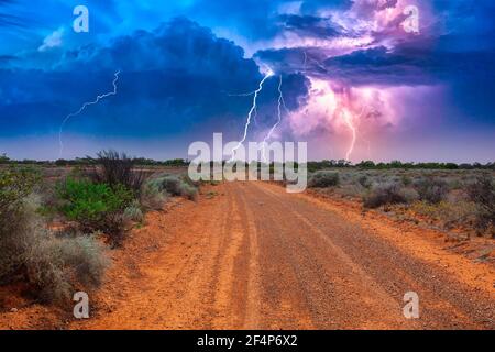 Verlassene australische Outback-Landschaft mit roter Schotterstraße zum Horizont Mit Büschen an Straßenrändern und schwerem Gewitter mit weißem Purpur Blitze Stockfoto