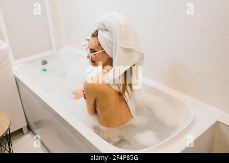 Im Badezimmer sitzt eine reizende Frau mit schneeweißem Lächeln. Stilvolle Frau in Handtuch auf dem Kopf und Sonnenbrille nimmt Spa-Behandlungen zu Hause oder im Hotel Stockfoto