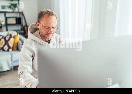 Lächelnder Mann mittleren Alters in Brillen mit einem modernen Computer in seinem Wohnzimmer. Schreiben oder Distanz oder freiberufliche Arbeit auf weltweite Quarantäne Zeit c Stockfoto