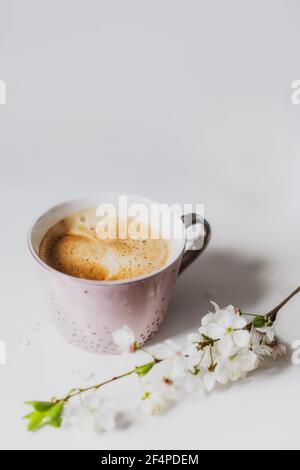 Eine Tasse Kaffee in einer hellrosa Tasse und Frühling blühenden Zweig mit Blumen auf dem weißen Hintergrund. Minimalist Vertikale Karte. Weicher, selektiver Fokus Stockfoto