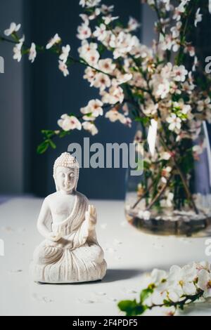 Dekorative weiße Buddha Statuette mit blutigen Ästen in der Vase auf dunklem Hintergrund. Meditations- und Entspannungsritual. Buddha Geburtstag Stockfoto