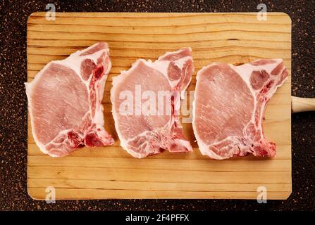 Draufsicht auf drei rohe Schweinekoteletts auf einem hölzernen Schneidebrett Stockfoto