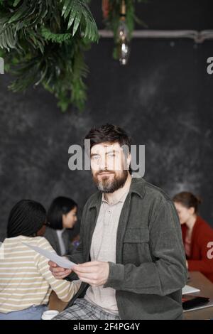 Vertikale Taille bis Porträt von zeitgenössischen bärtigen Geschäftsmann Blick auf Kamera mit verschiedenen Gruppen von Menschen treffen im Hintergrund Stockfoto