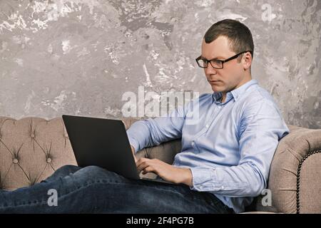 Ein junger erfolgreicher Mann in Hemd und Brille arbeitet mit einem Laptop, der auf einem Sofa im Home Office vor dem Hintergrund einer Betonwand liegt. Die CO Stockfoto