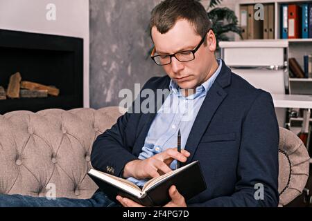 Junge erfolgreiche Mann in Anzug und Brille arbeitet mit Notizblock sitzen in großen geräumigen Home Office auf Sofa vor dem Hintergrund des Kamins. Kontra Stockfoto
