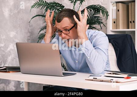 Porträt eines empörten Mannes in einem Hemd und Brille, Blick auf den Laptop-Bildschirm mit den Händen nach oben, schreien nach dem Monitor, sitzen im Büro ein Stockfoto