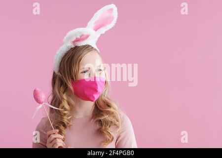 Frohe Ostern. stilvolles Kind mit langen welligen blonden Haaren mit rosa Maske, Hasenohren und osterei isoliert auf rosa Hintergrund. Stockfoto