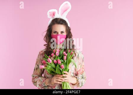 Portrait der modernen Frau im floralen Kleid mit Tulpen-Bouquet, Hasenohren und rosa Maske auf rosa Hintergrund. Stockfoto