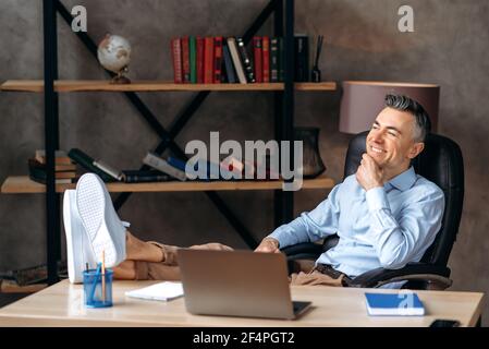 Zufrieden attraktiver grauhaariger Geschäftsmann, ceo oder Manager, am Arbeitsplatz ausruhen, seine Beine auf den Tisch werfen, sein Kinn mit der Hand halten, aus dem Fenster schauen, von einem Urlaub träumen, lächelt Stockfoto