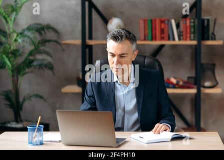 Erfolgreiche seriöse Erwachsene attraktive kaukasischen Business Leader oder ceo, sitzen am Arbeitsplatz im Büro in einem formalen Anzug, konzentriert arbeiten in einem Laptop, Surfen im Internet, analysiert Informationen Stockfoto