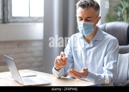 Vorsichtsmaßnahmen, Gesundheitskonzept. Ein erfolgreicher, reifer Büroangestellter in formeller Kleidung, in einer schützenden medizinischen Maske auf seinem Gesicht, sitzt an seinem Schreibtisch im Büro, desinfiziert seine Hände mit einem Antiseptikum Stockfoto