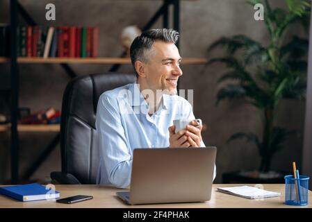 Fröhliche grauhaarige erfolgreiche attraktive Mann, Unternehmer oder Mitarbeiter, nahm eine Pause, sitzt an einem Schreibtisch, hält eine Tasse Kaffee, schaut zur Seite, Träume von einem Urlaub, lächelt Stockfoto