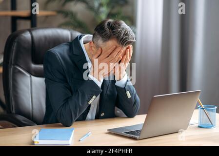 Erschöpfter kaukasischer grauhaariger Geschäftsleiter, Manager, ceo in formellem Anzug sitzt im Büro, er hat Kopfschmerzen, müde oder gestresst, bedeckt sein Gesicht mit den Händen, braucht Ruhe oder Pause Stockfoto