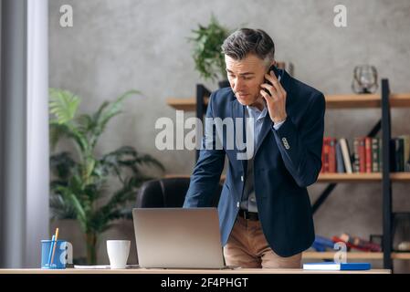 Vielbeschäftigter erfolgreicher, schöner grauhaariger kaukasischer Geschäftsmann, ceo, steht in formellem Anzug in seinem modernen Büro, spricht auf einem Mobiltelefon mit einem Kollegen oder Kunden, nutzt einen Laptop und im Internet surfen Stockfoto