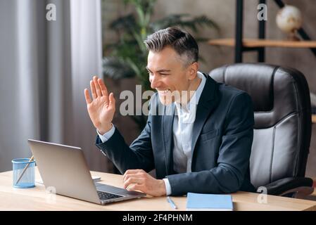 Gut aussehende erfolgreiche selbstbewusste reifen Geschäftsleiter oder Manager, in formellen Anzug, verwendet Laptop, kommuniziert per Videoanruf, grüßt Kollegen während Online-Meeting, winkt seine Hand, lächelt freundlich Stockfoto