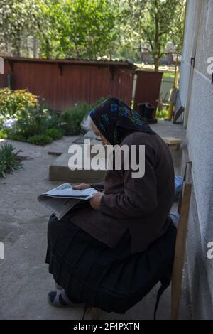 Sehr alte ältere Frau, die auf einem Stuhl im Hinterhof sitzt und Zeitung lesen Stockfoto