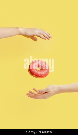 Donut fällt zwischen den Händen auf hellgelben Papierhintergrund. Donuts Lieferung, Ernährung, Zucker Konzept. Weibliche Hände und glasierte Donut mit Streuseln fliegen in der Luft. Vertikal. Kreatives Stillleben Stockfoto