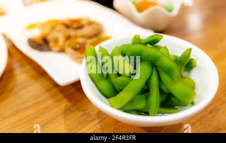 Edamame oder grüne Sojabohnen in weißer Schale auf Holztisch im japanischen Restaurant blanchiert. Schoten von grünen Sojabohnen auf verschwommenen japanischen Lebensmitteln. Hohe Faser Stockfoto