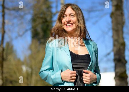 Glückliche Frau mittleren Alters in einem stilvollen blauen Jacke mit einem Fröhliches freundliches Lächeln in einer Nahaufnahme aus dem unteren Winkel Im Frühling im Freien in einem Park Stockfoto