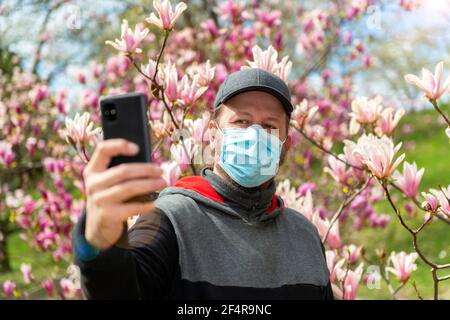 Ein junger Mann in einer medizinischen Maske macht Selfie mit Ein Smartphone in der Nähe von schönen Magnolien blüht während eines Lockdown Stockfoto