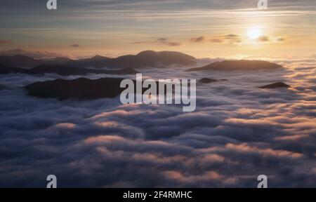 Sonnenuntergang versteckt hinter Wolken und Nebel über Hügeln, leuchtend gelbe Sonne auf bunte Wolkenlandschaft, blau violett orange Himmel. Stockfoto