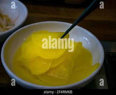 Hand mit Essstäbchen Pick gelb eingelegten Daikon Rettich in einer weißen Tasse zum Essen. Asiatische Beilage eingelegter Rettich, koreanische Küche traditionell. Selektiv Stockfoto