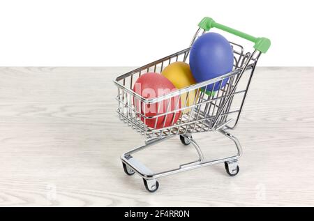 Drei farbige Eier in einem Mini-Einkaufswagen auf einem Weißer Holztisch auf weißem Hintergrund Stockfoto