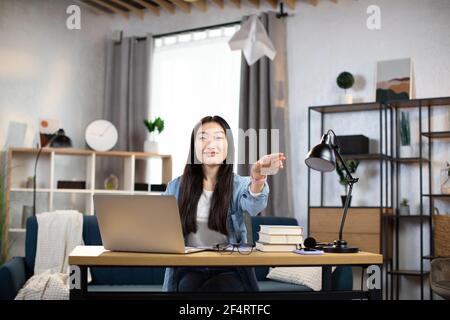 Lächelnde Freiberuflerin, die mit Papierflugzeug spielt, während sie von zu Hause aus arbeitet. Attraktive asiatische Frau, die Pause während der Arbeitszeit. Stockfoto