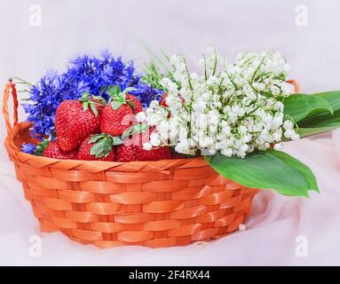 Hübsche Lilien des Tals, rote frische Erdbeeren und Kornblumen in orangefarbenem Korb Stockfoto