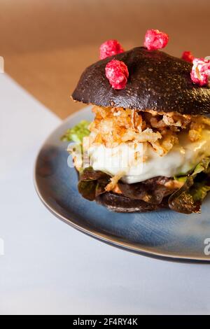 Corona (antiCOVID) Cheeseburger mit hausgemachten schwarzen Brötchen, Salat, gebratene Zwiebel und Käse auf einem Teller auf einem Backpapier. Restaurant, Burger Bar. Stockfoto