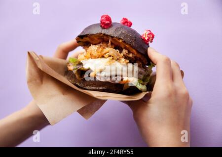 Corona (antiCOVID) Cheeseburger mit hausgemachten schwarzen Brötchen, Salat, gebratene Zwiebel und Käse in weiblichen Händen auf violettem Hintergrund. Restaurant, Burger Bar Stockfoto