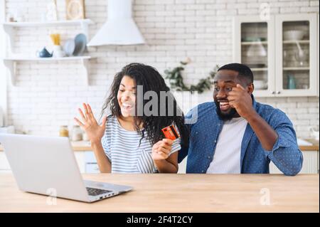 Überglücklich junge multirassische Paar mit einer Kreditkarte zu machen Kauf online. Fröhliche afroamerikanische Männer und Frauen genießen den gemeinsamen Einkauf zu Hause und feiern den lang ersehnten Kauf Stockfoto