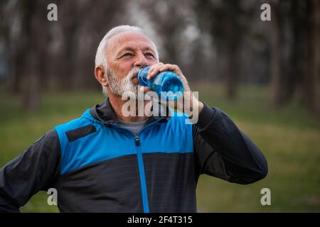 Der ältere Mann trinkt Wasser, nachdem er im Park trainiert hat. Stockfoto