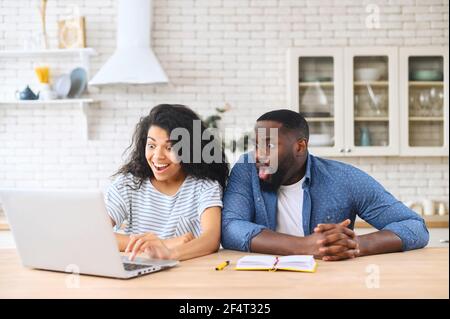 Schockiert multirassischen Paar in der Liebe starrte auf den Laptop-Bildschirm, überrascht mit unerwarteten Nachrichten oder E-Mail schaut auf den Computer sitzen am Tisch in der Küche zu Hause Stockfoto