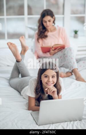 Tochter beschäftigt am Laptop, Mutter liest hinter. Stockfoto
