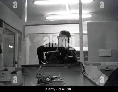 1964, historisch, in einem abgeteilten Büro in einem Industrieunternehmen, ein männlicher Mitarbeiter, der an einer Theke oder einer Luke steht und ein Gespräch auf einem großen Bakerlite-Telefon führt, England, Großbritannien. Auf dem Bild ist eine Schreibmaschine zu sehen. Die Brtish Firma Wolseley Webb, hergestellt unter Lizenz, der Gartenrotator, der Merry Tiller, zu der Zeit, als britische Technik mit Qualität gleichgesetzt und für die Dauer gebaut wurde. Stockfoto