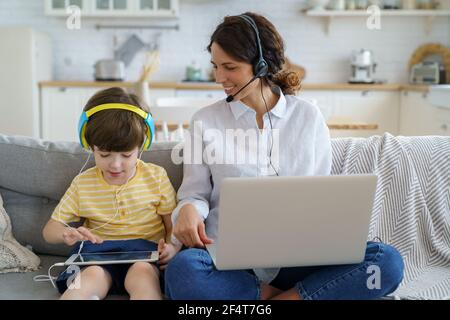 Glückliche Mutter mit Kind sitzt auf dem Sofa zu Hause während Lockdown Arbeit auf Laptop Kind spielen im Tablet Stockfoto