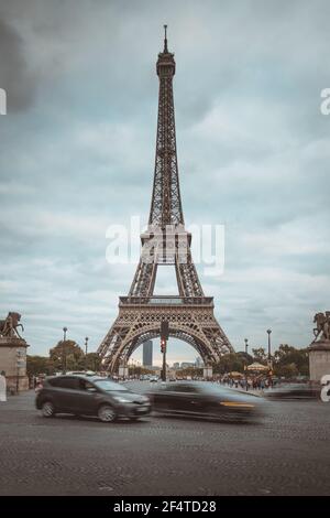 Autos, die an einem bewölkten Tag am Eiffelturm in Paris vorbeifahren. Langzeitbelichtungen zur Anzeige der Bewegung der Fahrzeuge an der Ampel. Stockfoto