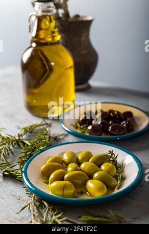 Natürliche schwarze und grüne Oliven mit Rosmarin-, Basilikum- und Olivenbaumblättern auf blauem Holzhintergrund. Stockfoto