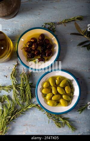 Natürliche schwarze und grüne Oliven mit Rosmarin-, Basilikum- und Olivenbaumblättern auf blauem Holzhintergrund. Draufsicht. Stockfoto