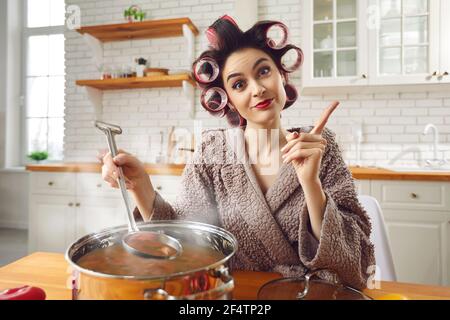Junge lustige attraktive Hausfrau in Lockenwickler und Bademantel Kochen an Wohnküche Stockfoto