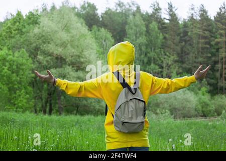 Eine junge Frau in einem gelben Hoodie mit erhobenen Händen steht im Wald zurück. Offen für eine Reise. Stockfoto