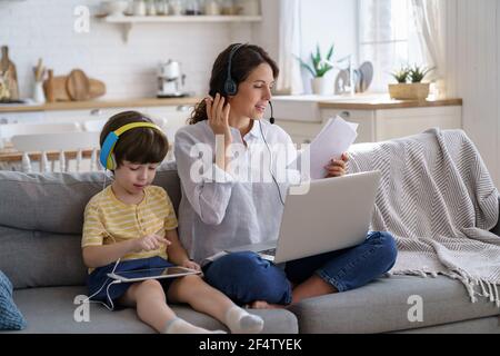 Freiberufliche Mutter sitzt auf dem Sofa im Home Office während Lockdown Arbeit auf Laptop, Kind spielt auf dem Tablet Stockfoto