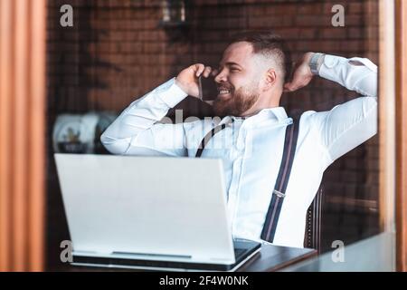 Junger Hipster, Geschäftsmann, der in einem Café telefoniert, Laptop auf dem Tisch Stockfoto